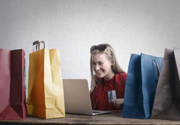 A joyful woman shopping online using a laptop with colorful shopping bags surrounding her.