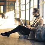 Man sitting by a window at a Budapest train station, working on his laptop with sunlight streaming through.